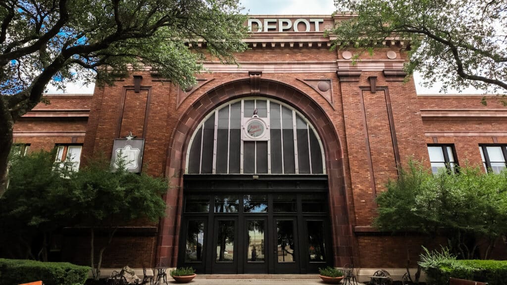 The iconic Katy Depot — where history and elegance meet. Front entrance of the historic Katy Depot building, featuring a grand arched window, red brick façade, and decorative stone details, framed by large leafy trees.